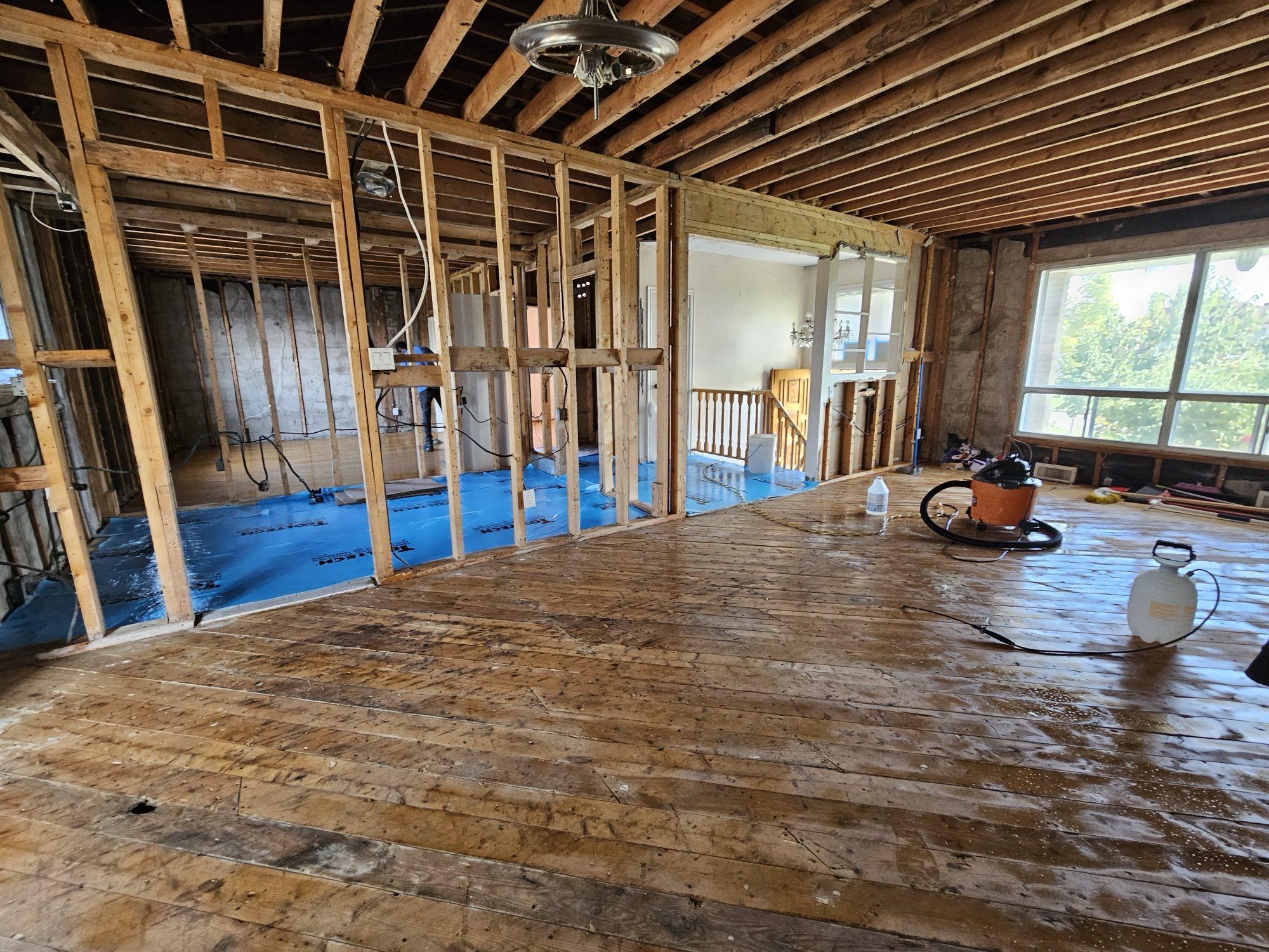 A main floor stripped to the studs and joists, with the original plank subfloor exposed and blue vapour barrier laid in the next room