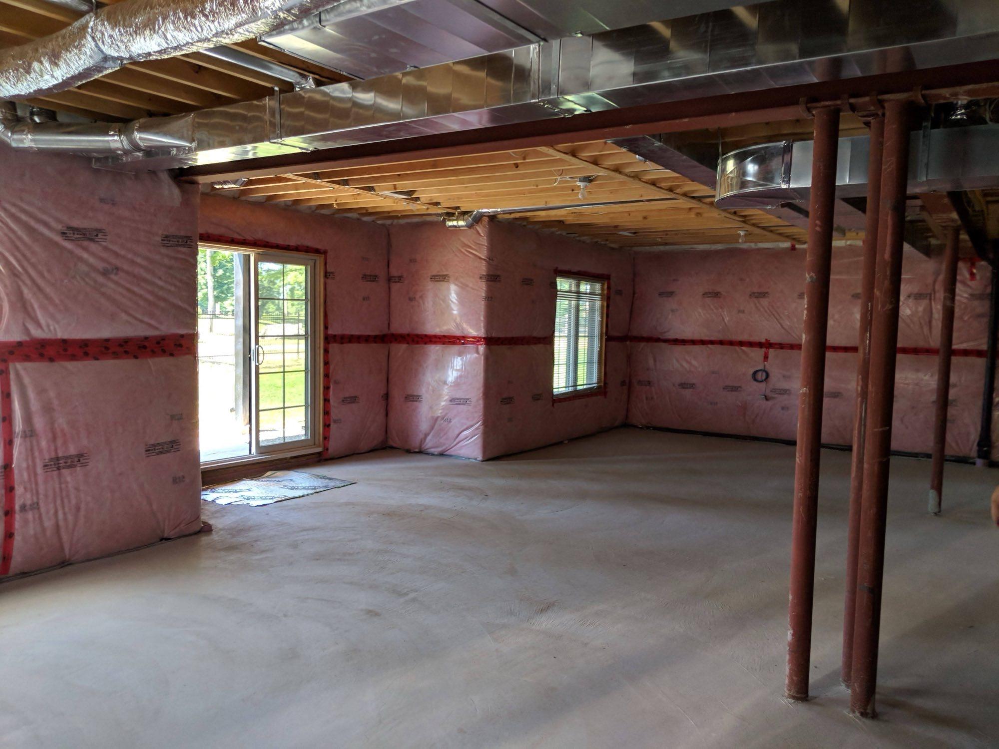 A basement at rough-in: pink batt insulation taped along every wall, new ductwork run under the joists, steel posts and a bare concrete slab